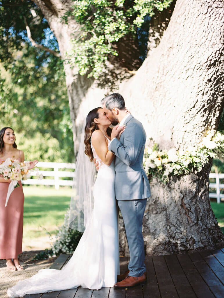wedding ceremony at circle oak ranch, the bride and groom having their first kiss in front of a 500 year old oak tree while their bridal party happily cheers