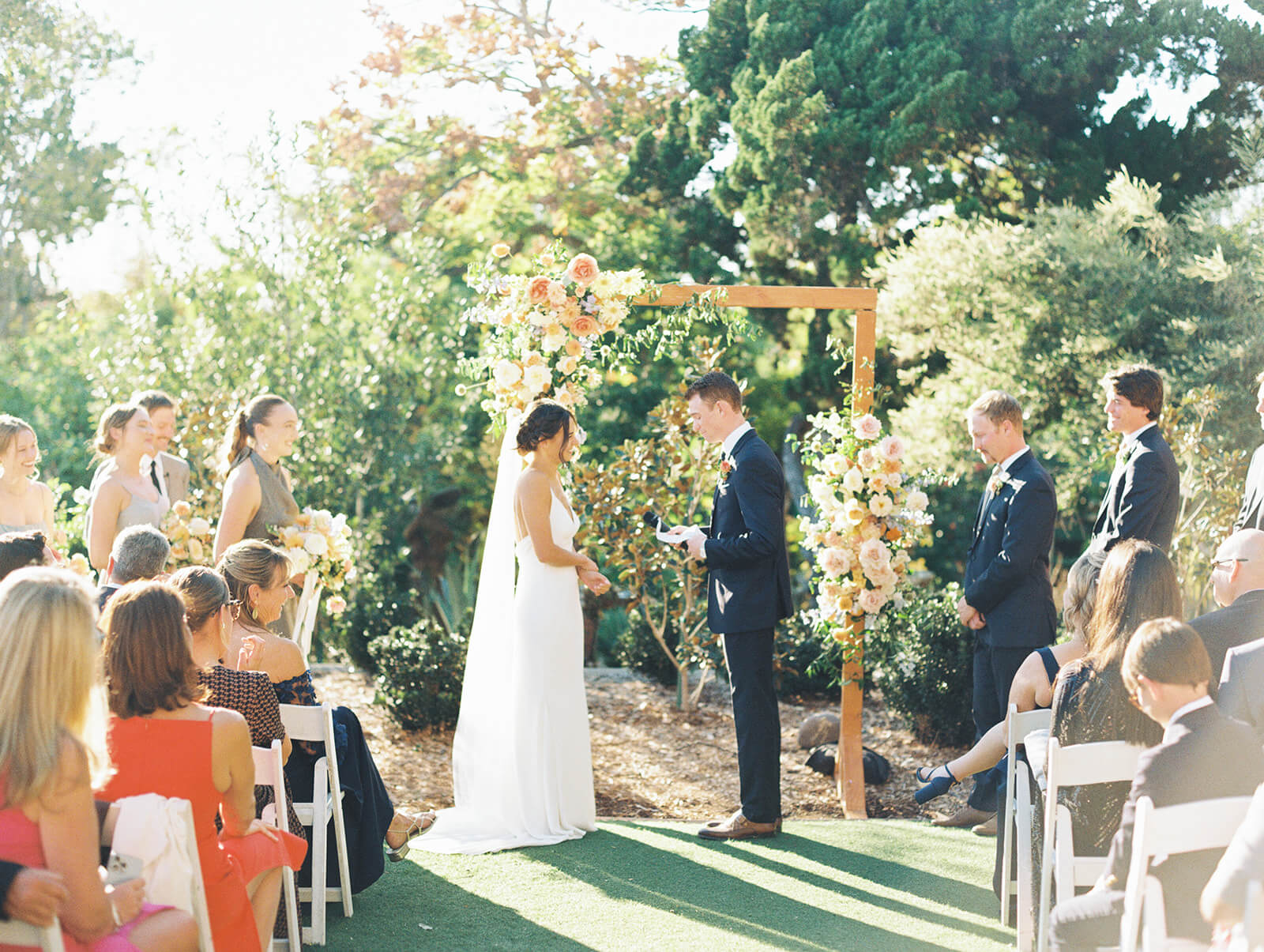 A groom standing at a wedding ceremony alter reading his vows to his bride. there are flowers on the arch behind them, the wedding party is standing watching and guests are seated at chairs in front