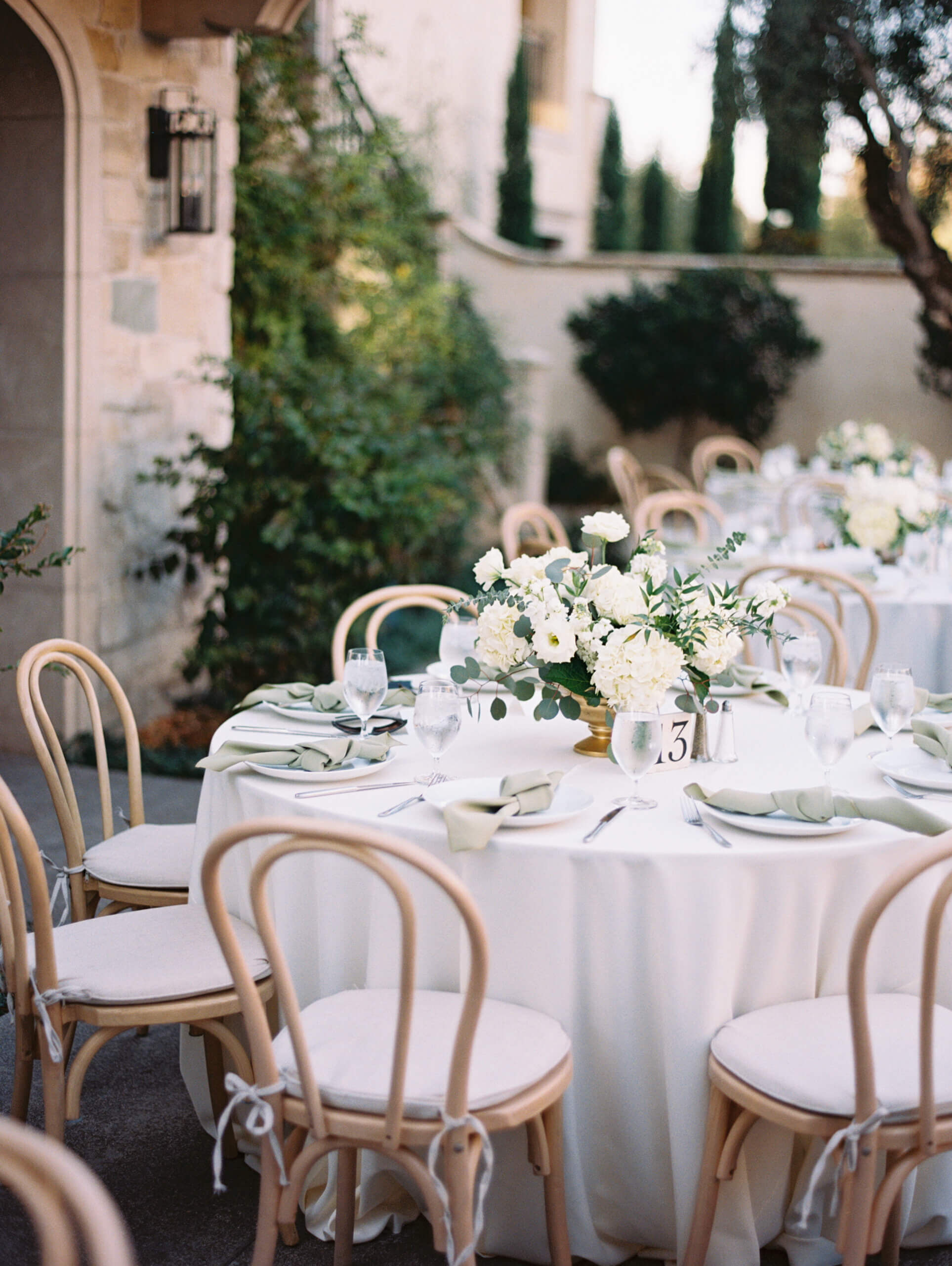 a wedding day reception table with european architectural features in the background, on the table is a white and green floral arrangement in a gold vase, light wood cross back chairs with white cushions around the round tables