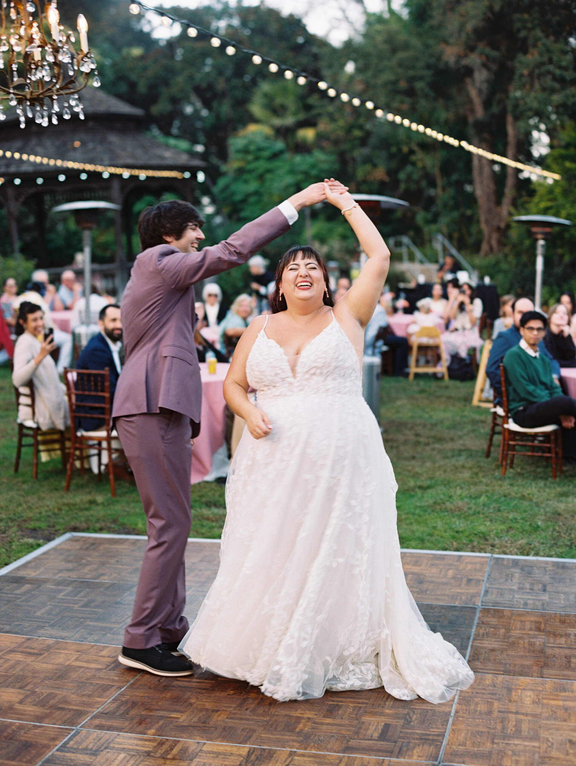 groom wearing purple suit spins laughing bride wearing ivory wedding gown during their first dance under market lights at San Diego Botanic Garden